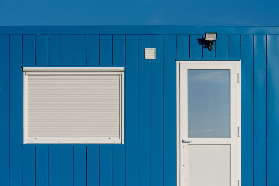 Close-up of a blue building facade featuring a white door and window. Minimalist architecture.
