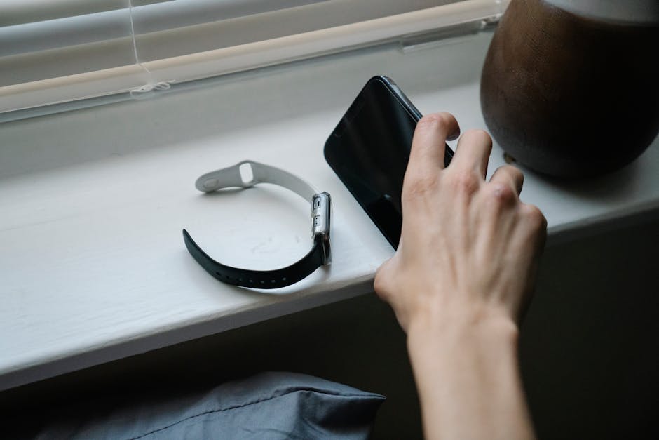 From above of crop anonymous person taking cellphone placed on windowsill near smart watch while waking up in morning