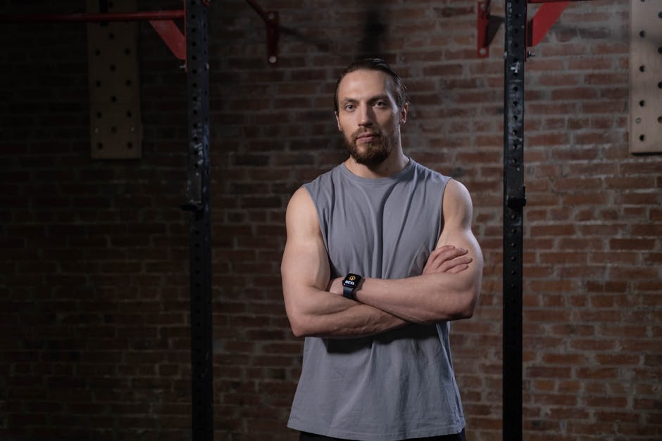 A muscular man in a gray tank top stands with arms crossed in a gym setting.